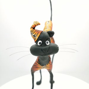 Könnte beinhalten: Eine schwarze Katzenfigur mit orange-schwarzen Tigerstreifen, die auf einer weißen Oberfläche steht. Die Katze hat einen langen, dünnen Schwanz und schaut direkt in die Kamera.