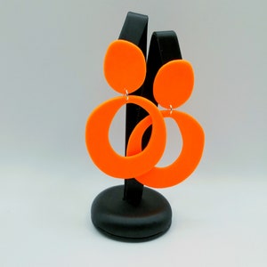 May include: A pair of orange statement earrings with a large circle and a smaller circle on top. The earrings are displayed on a black stand.