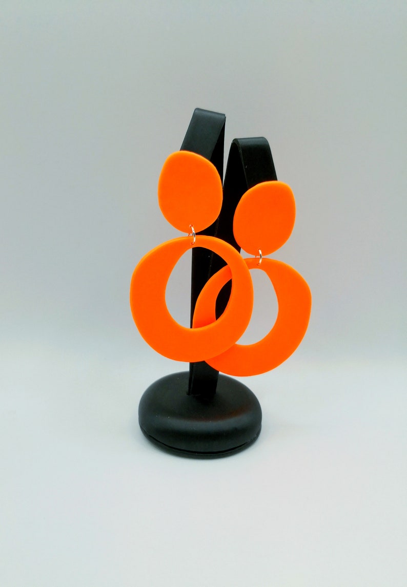 May include: A pair of orange statement earrings with a large circle and a smaller circle on top. The earrings are displayed on a black stand.