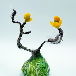 Könnte beinhalten: Eine kleine Keramikvase mit grünem und weißem Muster, verziert mit einem gedrehten Zweig mit zwei gelben Blüten.