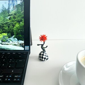 Könnte beinhalten: Eine kleine, rote, stachelige Blumenskulptur auf einem schwarz-weiß karierten Sockel. Die Skulptur steht auf einer weißen Oberfläche neben einem Laptop und einer Tasse Kaffee.