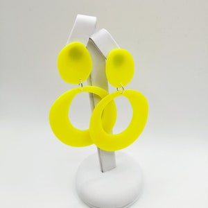 May include: A pair of bright yellow statement earrings with a large oval shape and a smaller round top. The earrings are displayed on a white stand.