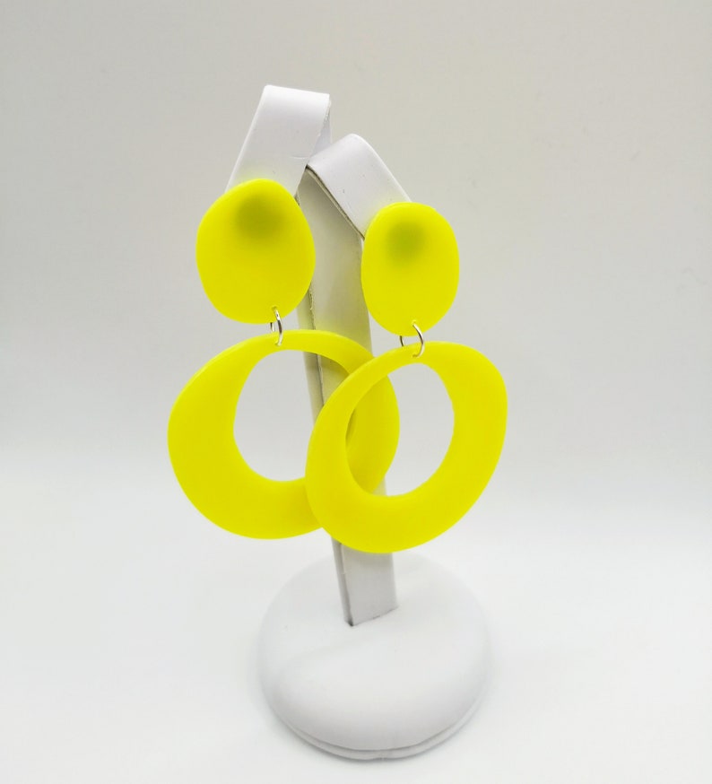 May include: A pair of bright yellow statement earrings with a large oval shape and a smaller round top. The earrings are displayed on a white stand.