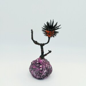Könnte beinhalten: Eine kleine Keramikskulptur eines Baumes mit einer einzigen orangefarbenen Blüte. Der Baum besteht aus schwarzem Draht und die Blüte aus orangefarbenem und schwarzem Ton. Die Skulptur steht auf einem violetten und weißen Keramiksockel.