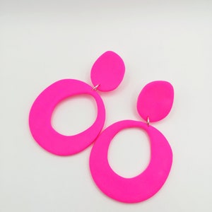 May include: A pair of neon pink statement earrings with a large oval shape and a smaller oval accent. The earrings are made of a smooth, matte material.