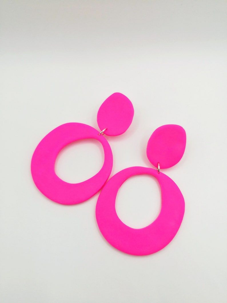 May include: A pair of neon pink statement earrings with a large oval shape and a smaller oval accent. The earrings are made of a smooth, matte material.