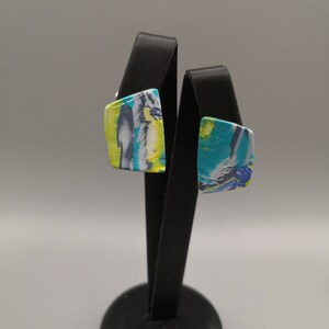 May include: A pair of square-shaped earrings with a colourful abstract design. The earrings are displayed on a black stand.