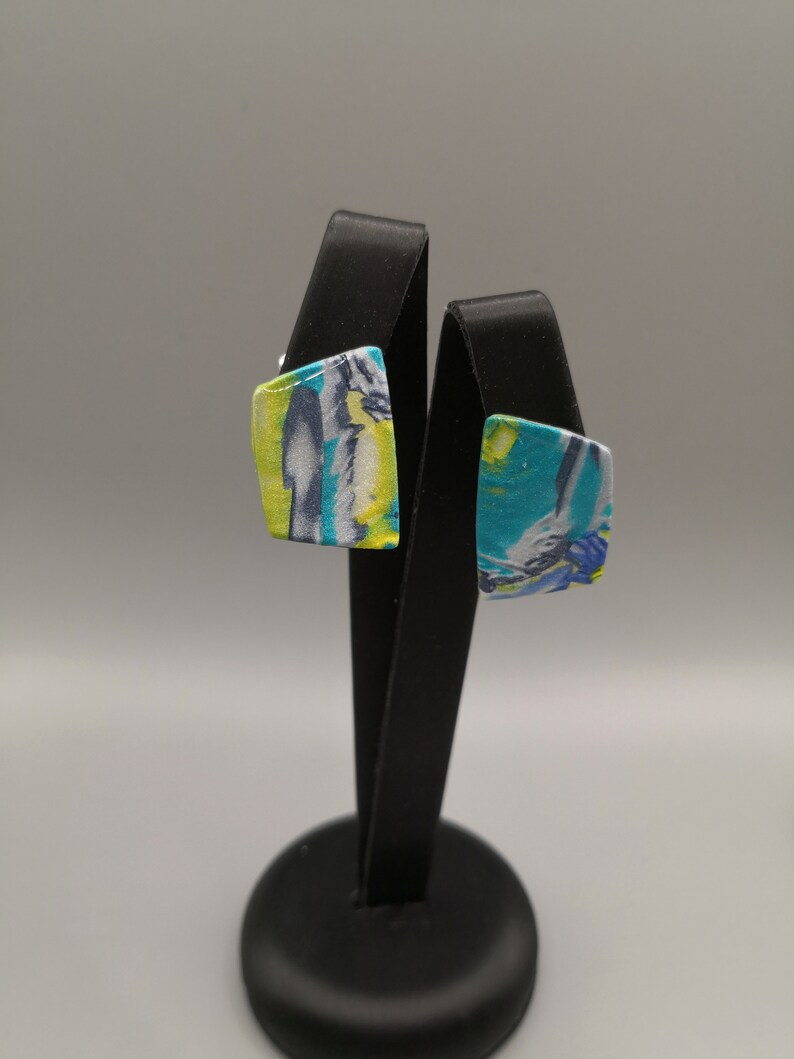 May include: A pair of square-shaped earrings with a colourful abstract design. The earrings are displayed on a black stand.