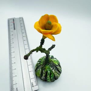 Könnte beinhalten: Eine kleine, gelbe Blume mit grünem Zentrum auf einem grün-braunen Sockel. Die Blume ist aus Polymer Clay gefertigt und etwa 5 cm hoch.