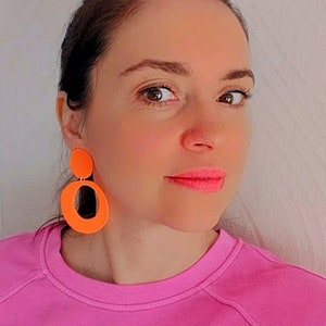 May include: A woman wearing a pink sweatshirt with bright orange geometric earrings. The earrings are large and dangle from her ears.
