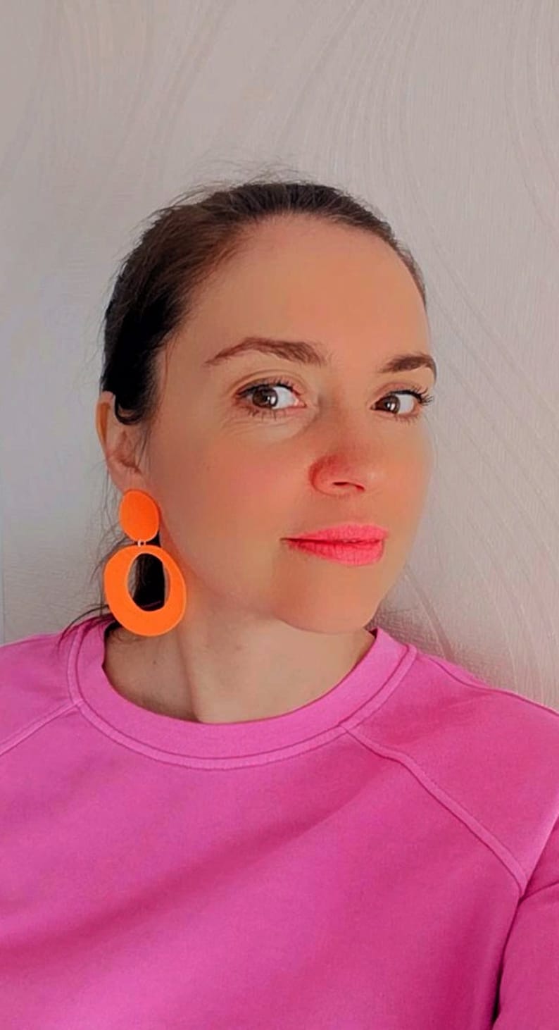 May include: A woman wearing a pink sweatshirt with bright orange geometric earrings. The earrings are large and dangle from her ears.