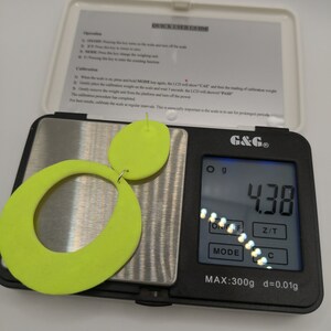 May include: A digital scale with a neon green pair of earrings on the platform. The scale reads 4.38 grams. The scale has a maximum weight capacity of 300 grams and a precision of 0.01 grams.