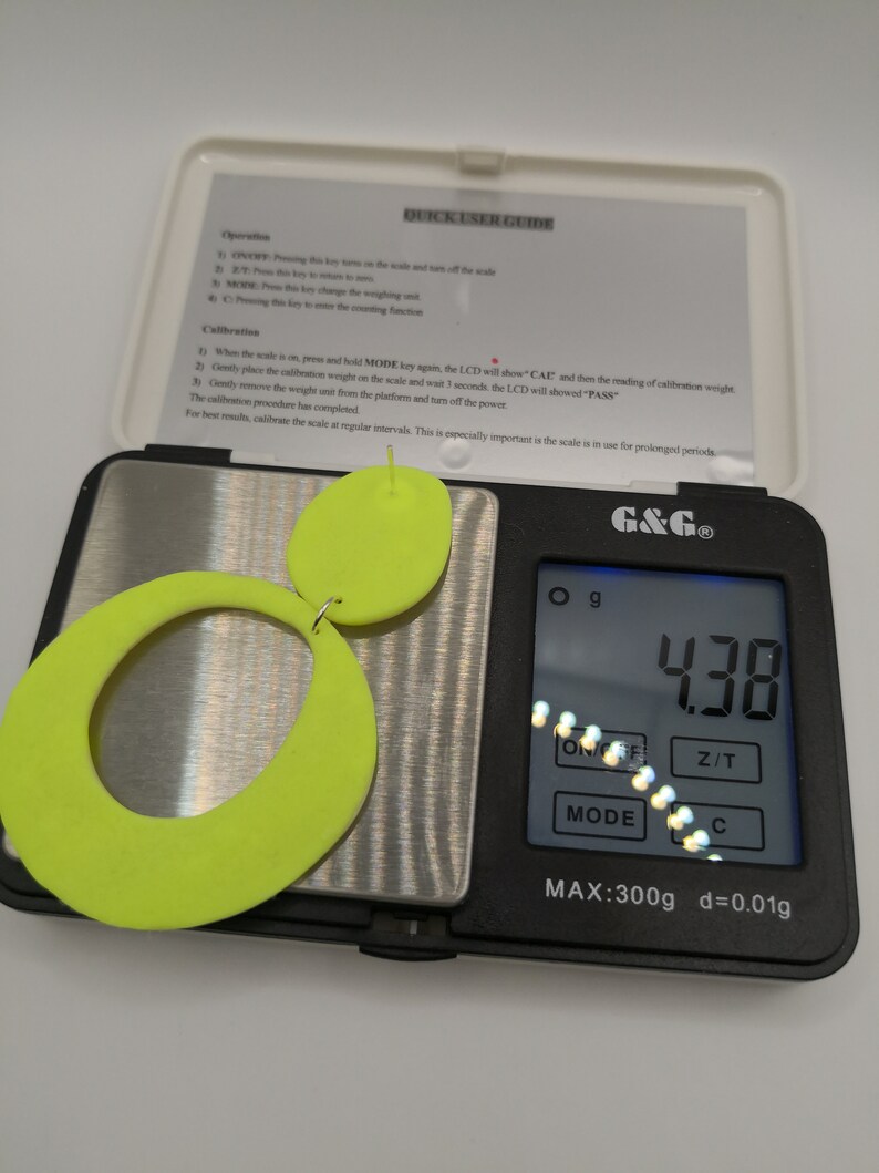 May include: A digital scale with a neon green pair of earrings on the platform. The scale reads 4.38 grams. The scale has a maximum weight capacity of 300 grams and a precision of 0.01 grams.