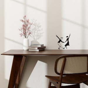 Peut inclure: Une décoration murale en métal noir représentant une silhouette de couple dansant. La décoration est fixée sur un mur blanc au-dessus d'un bureau en bois avec une chaise.