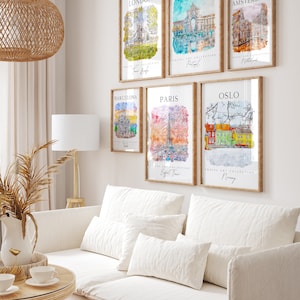 May include: A white wall with six framed watercolor paintings of famous landmarks in different cities. The paintings are in wooden frames with a light brown finish. The cities featured are London, Lisbon, Amsterdam, Barcelona, Paris, and Oslo. The text on the paintings includes the city name, the country, and the name of a landmark in the city.