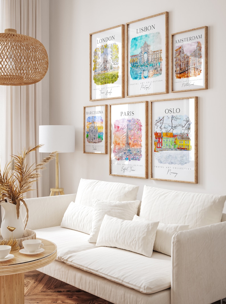 May include: A white wall with six framed watercolor paintings of famous landmarks in different cities. The paintings are in wooden frames with a light brown finish. The cities featured are London, Lisbon, Amsterdam, Barcelona, Paris, and Oslo. The text on the paintings includes the city name, the country, and the name of a landmark in the city.