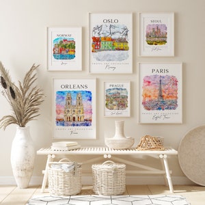 May include: Six watercolor paintings of different cities around the world, including Oslo, Norway, Seoul, Paris, Orleans, and Prague. Each painting is framed in a white frame and hangs on a white wall. The paintings are arranged in a grid pattern. The wall is decorated with a vase of dried flowers and a white bench with two wicker baskets.