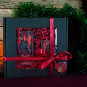 Op de afbeelding: Een zwarte geschenkdoos met een venster, vastgebonden met een rood lint en strik. Binnenin omringt rood gescheurd papier een kaart met een figuur en het woord "SLICK". De doos is versierd met kerstgroen.
