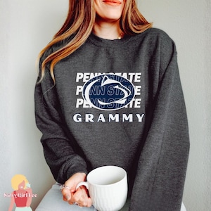 May include: A dark gray sweatshirt with the text "PENNSYLVANIA STATE" stacked vertically, with the Penn State logo in the center. The word "GRAMMY" is below the logo. A white mug is held in the person's hands.