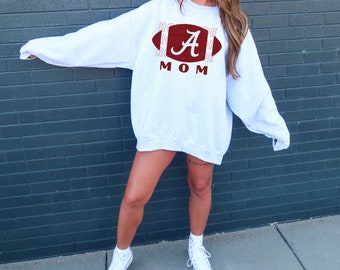 May include: A light gray sweatshirt with a maroon football graphic and the word "A" in white script. Below the graphic, the word "MOM" is printed in maroon. The word "ROSE" is printed on the left and right sides of the football graphic.