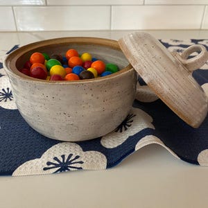 May include: A white ceramic bowl with a lid, filled with colorful candy, sitting on a blue and white floral patterned fabric.