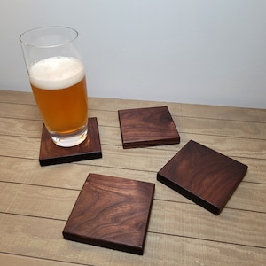 May include: Four dark brown wooden coasters with a glass of beer on one of them. The coasters are square and have a smooth surface.