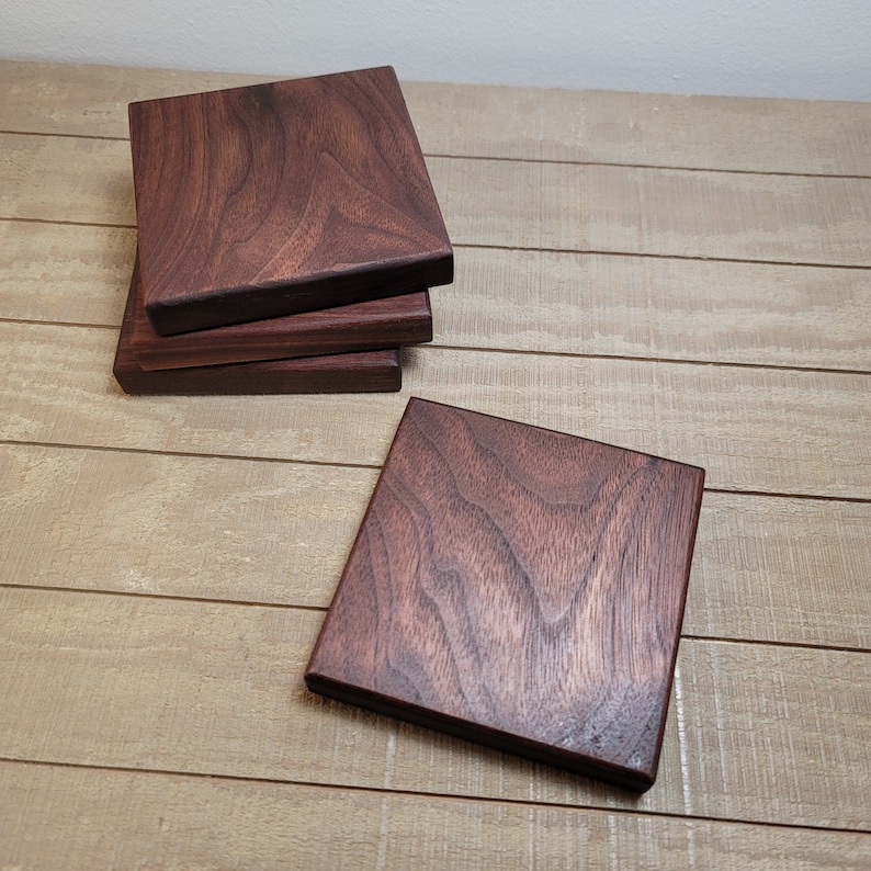 May include: Four square wooden coasters made of dark walnut wood. The coasters are stacked on top of each other, with the top coaster showing a natural wood grain pattern.