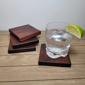 May include: Four dark brown wooden coasters stacked on top of each other. A glass of water with a lime wedge sits on top of the coasters.