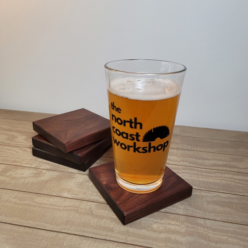 May include: Four dark brown wooden coasters with a glass of beer on top. The glass has a logo that says "the north coast workshop".
