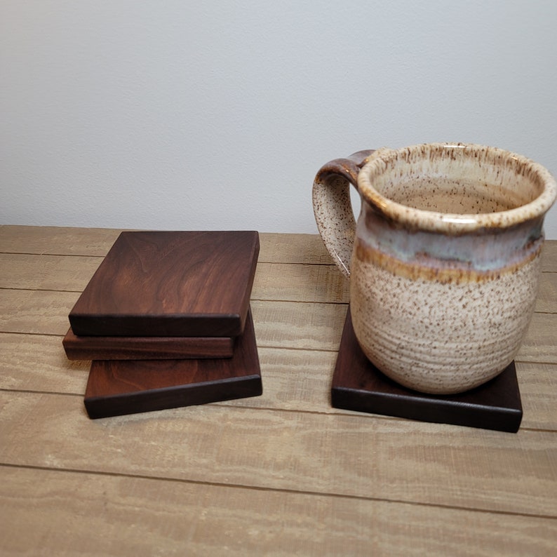 May include: Four dark brown wooden coasters stacked on a wooden surface. A speckled brown and white ceramic mug sits on a single coaster.