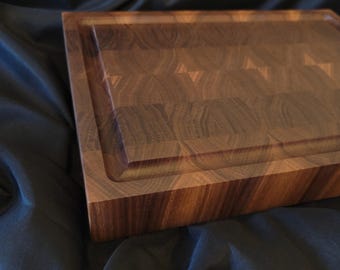 May include: A large, rectangular, wooden cutting board made of walnut wood. The board has a raised edge and a checkered pattern.
