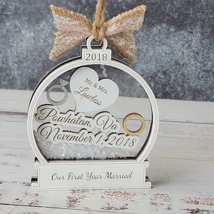 May include: A white wooden ornament with a clear center, featuring a heart, rings, and the text "Mr. & Mrs. Lawless." It includes the date "2018" and the text "Powhatan, Va November 1, 2018." The base reads "Our First Year Married."
