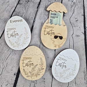 May include: Four wooden Easter egg decorations. Two are white, two are natural wood. Each egg has the words "Happy Easter" and a floral design. One egg is open, revealing a twenty-dollar bill. One egg has the name "Everest".