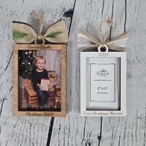 May include: Two wooden Christmas ornament picture frames. The frame on the left is brown with a burlap bow and green foliage. The frame on the right is white with a white lace bow. Both frames have a 2x3 inch picture opening. The left frame is engraved with "Everest Brooks" and "Christmas 2024". The right frame is engraved with "2024", "EverLee", "EverLeeCreations.com", "2" x 3" Picture Size", and "First Christmas Married".