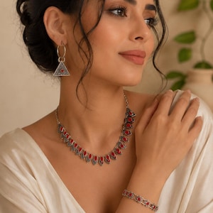 Peut inclure: Un ensemble de bijoux argentés composé d'un collier, d'un bracelet et de boucles d'oreilles créoles. Le collier et le bracelet présentent un motif répétitif de petites pierres rouges en forme de goutte serties dans du métal argenté oxydé.