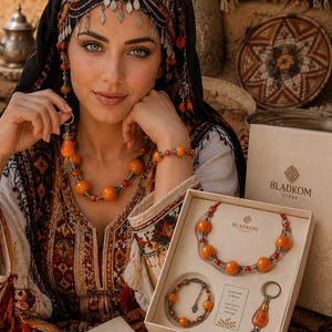 Peut inclure: Un coffret cadeau de bijoux dans une boîte beige comprenant un collier et un bracelet avec des perles orange et des détails argentés, ainsi qu'un porte-clés avec une perle orange et une breloque hamsa. La boîte porte le texte BLADKOM STORE.