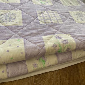 May include: A quilted blanket featuring a patchwork design of light purple and white fabric squares. The pattern includes alternating blocks of solid light purple, white with purple polka dots, white with purple floral prints, and white with purple plaid.