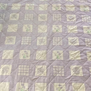 Op de afbeelding: Een rechthoekige quilt met een herhalend rasterpatroon van lichtpaarse stoffen vierkanten afgewisseld met gebroken witte vierkanten. De gebroken witte vierkanten tonen diverse bedrukte ontwerpen, waaronder bloemmotieven, ruitpatronen en kleine stippen.