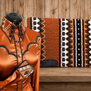 May include: A burnt orange long-sleeve shirt with a pointed collar, featuring black trim, silver studs, and white pearl accents. Beside it sits a rectangular saddle pad with a textured burnt orange, black, and white geometric pattern.