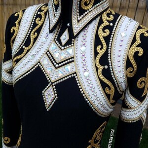 Könnte beinhalten: Ein schwarzes Langarmshirt mit weißen Einsätzen an Brust und Schultern, verziert mit goldener Schnörkelstickerei und zahlreichen irisierenden Kristallen. Der Kragen und der Brustbereich sind dicht mit klaren Strasssteinen besetzt.