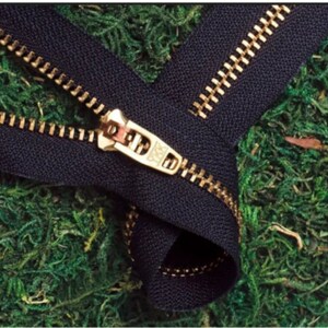 May include: Close-up of a black zipper with gold-colored teeth and a matching pull tab. The zipper is partially open, revealing the interlocking teeth. The background is a bed of green moss.