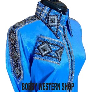 Könnte beinhalten: Ein leuchtend blaues Langarmhemd mit aufwendigen Verzierungen aus klaren, schwarzen und blauen Kristallen, die in geometrischen Rautenmustern an Kragen, Schultern und Manschetten angeordnet sind. Der Text BOBBY WESTERN SHOP ist schwarz aufgedruckt.