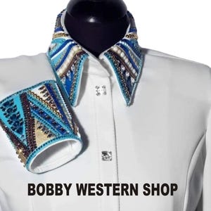 Könnte beinhalten: Weißes Westernhemd mit einem blauen und goldenen Perlenkragen und passenden Manschetten. Das Hemd hat einen Knopfleistenverschluss und einen spitzen Kragen. Der Text "BOBBY WESTERN SHOP" ist unten im Bild gedruckt.