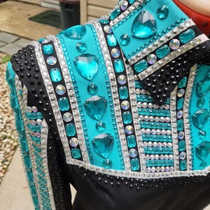 May include: A black and turquoise long-sleeve shirt featuring a high collar and intricate embellishments. The garment is heavily decorated with large, faceted turquoise crystals, smaller iridescent rhinestones, and rows of tiny black studs.