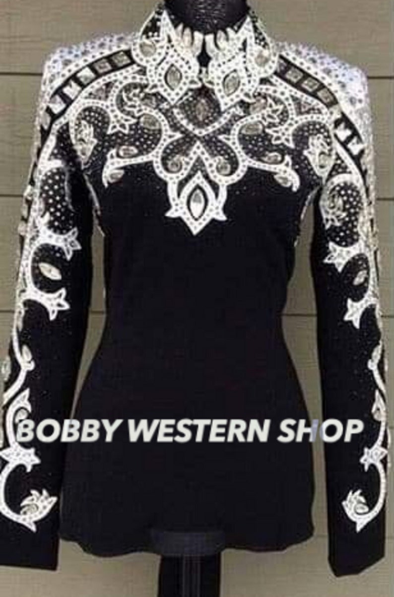 May include: Black long-sleeve shirt with intricate white and silver embellishments. The shirt features a high collar and detailed patterns on the shoulders and sleeves. The text "BOBBY WESTERN SHOP" is printed on the lower part of the shirt.