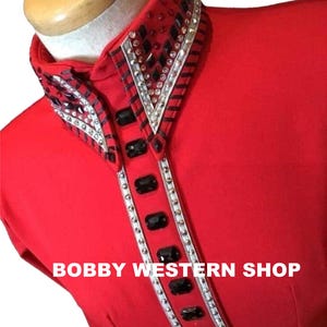 Könnte beinhalten: Ein leuchtend rotes Hemd mit hohem Kragen und einer vertikalen Knopfleiste, die mit Reihen aus klaren Kristallen und großen schwarzen rechteckigen Steinen verziert ist. Der Text BOBBY WESTERN SHOP ist in Weiß auf dem roten Stoff gedruckt.