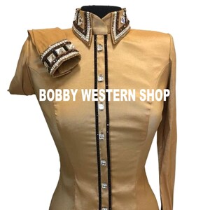 Könnte beinhalten: Ein goldfarbenes Langarmhemd mit hohem Kragen und Manschetten, die mit braunen und klaren Kristallen verziert sind. Die Knopfleiste hat einen schwarzen vertikalen Besatz mit quadratischen Kristallknöpfen. Der Text BOBBY WESTERN SHOP ist sichtbar.