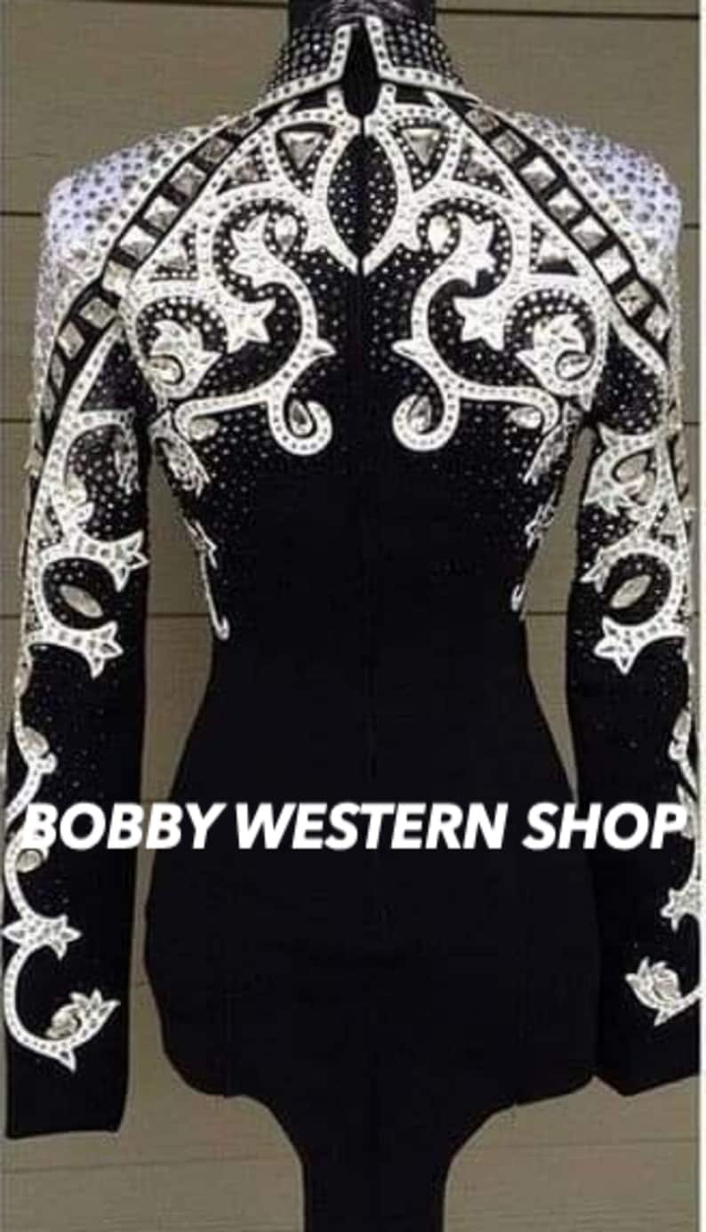 May include: Black equestrian show jacket with intricate white and silver embellishments. The jacket features a high collar and long sleeves, with ornate patterns of rhinestones and decorative elements. The text "BOBBY WESTERN SHOP" is visible at the bottom.