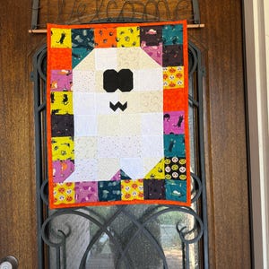 May include: A rectangular quilted wall hanging featuring a central white ghost figure with black eyes and a mouth. The ghost is surrounded by a border of colorful fabric squares patterned with black cats, orange pumpkins, and purple bats, finished with an orange trim.