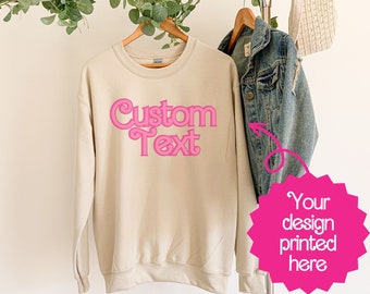 May include: A beige sweatshirt with the text "Custom Text" printed in pink on the front. The text is in a retro, stylized font.